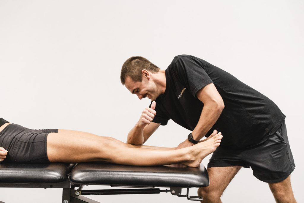A male physical therapist, wearing a black t-shirt and shorts, uses his elbow to apply pressure to the lower leg of a patient, who is lying on a treatment table. The therapist is smiling and looking down at the patient's leg.